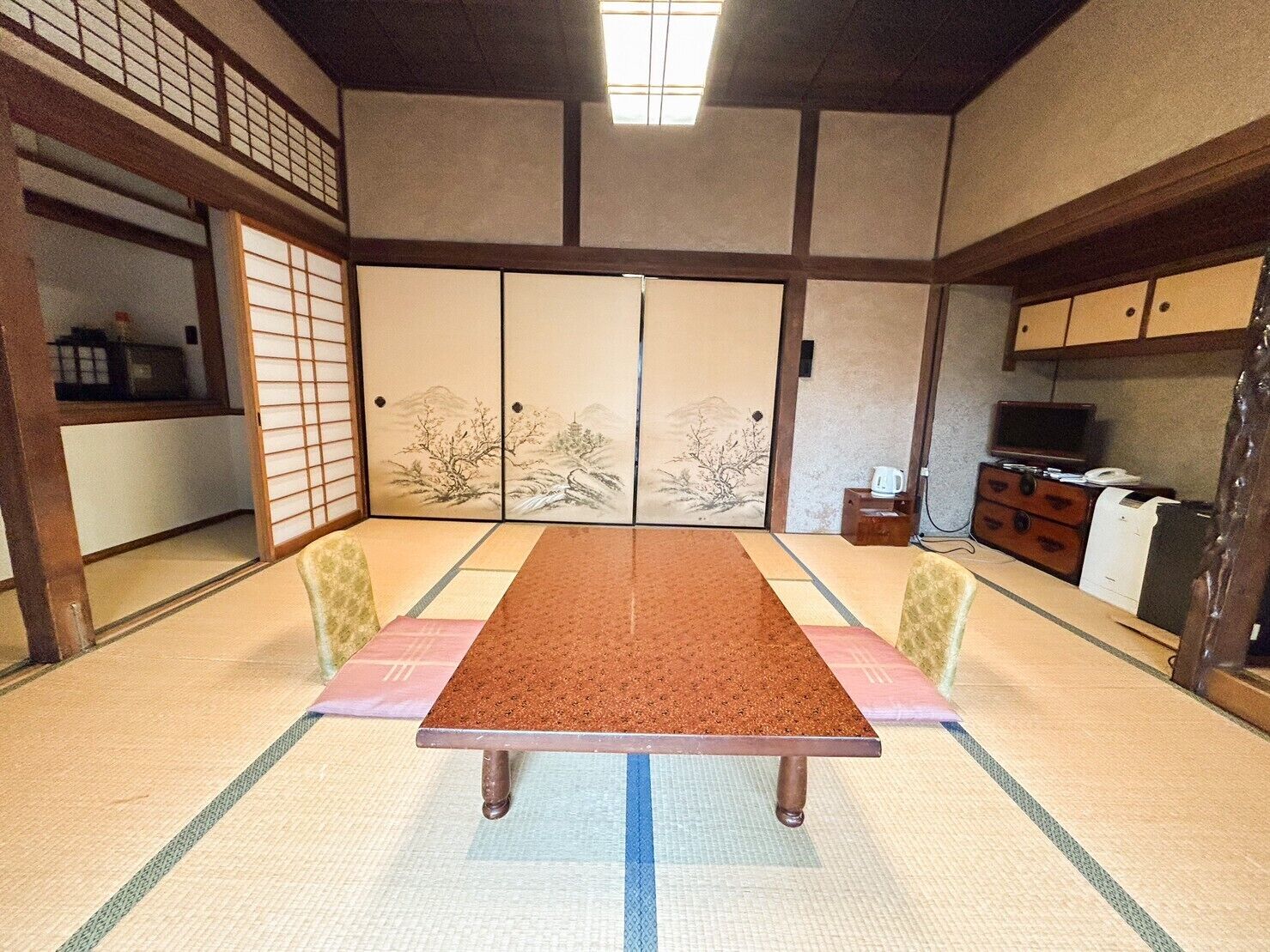 A traditional Japanese room with tatami flooring, a low table, sliding doors with artwork, and a television on a wooden cabinet.