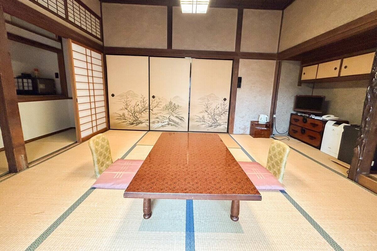 A traditional Japanese room with tatami flooring, a low table, sliding doors with artwork, and a television on a wooden cabinet.