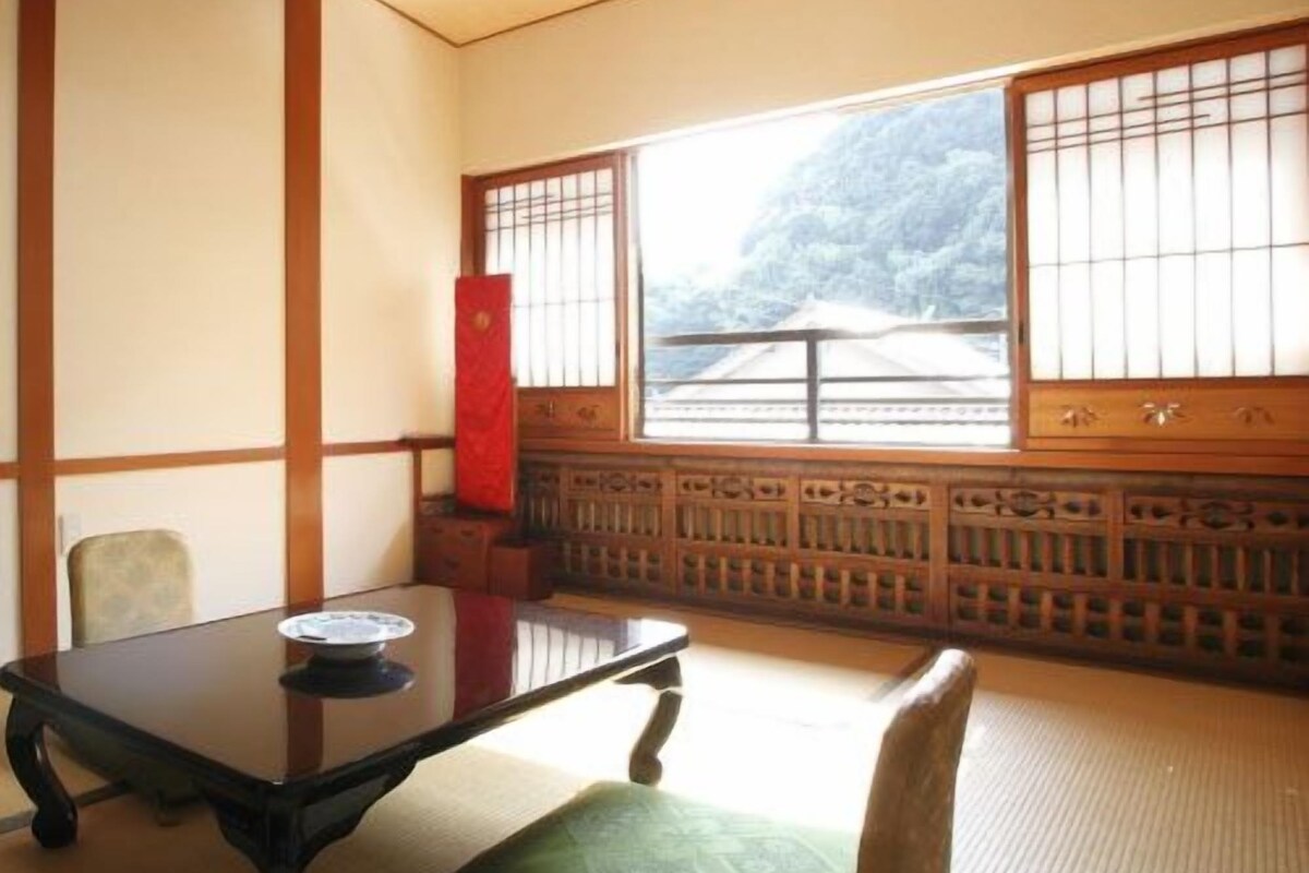 Una tradizionale stanza giapponese con pavimento in tatami, un tavolino basso, un cuscino e uno shoji che si apre su un paesaggio montano.