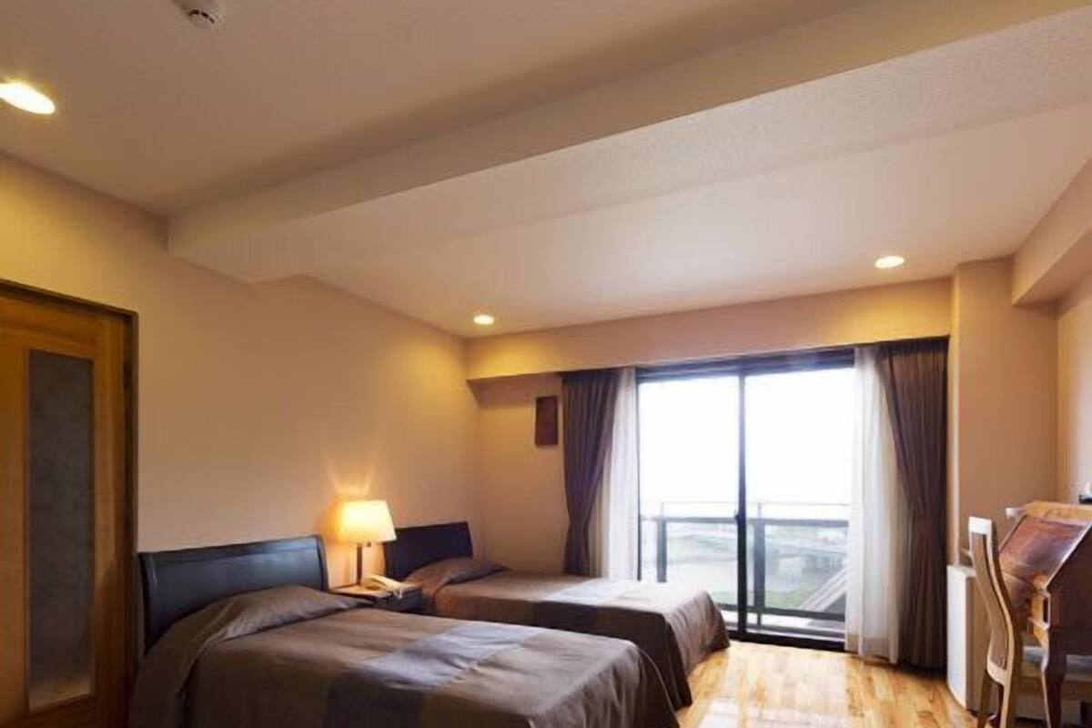 A hotel room with two beds, a desk, and a large window with curtains.