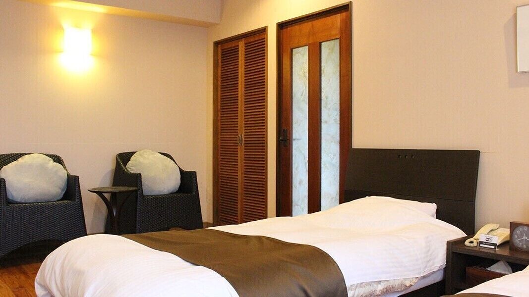 A hotel room with a bed, two chairs, a bedside table with a phone and a clock, and a wardrobe with a mirror.