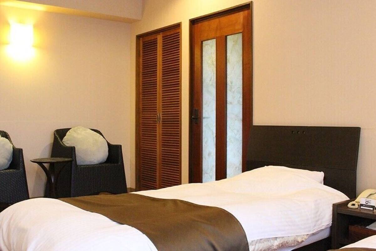 A hotel room with a bed, two chairs, a bedside table with a phone and a clock, and a wardrobe with a mirror.