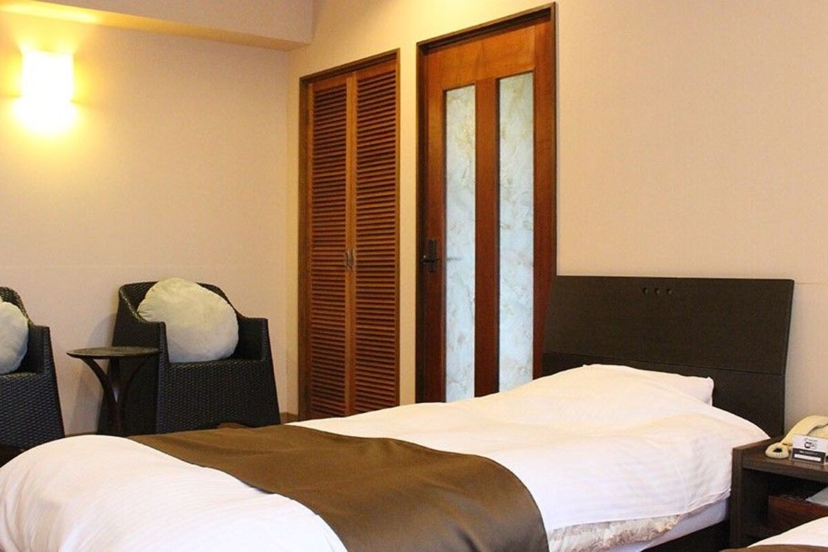 A hotel room with a bed, two chairs, a bedside table with a phone and a clock, and a wardrobe with a mirror.