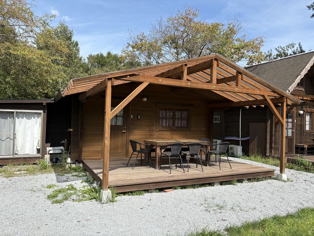 Eine Holzterrasse mit Tisch und Stühlen, ein überdachter Bereich mit Dach und ein kleines Gebäude mit einem Fenster.