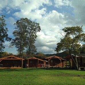 A row of wooden cabins with a grassy lawn in front, large trees, and a cloudy sky. - Pension Himawari (Karuizawa)