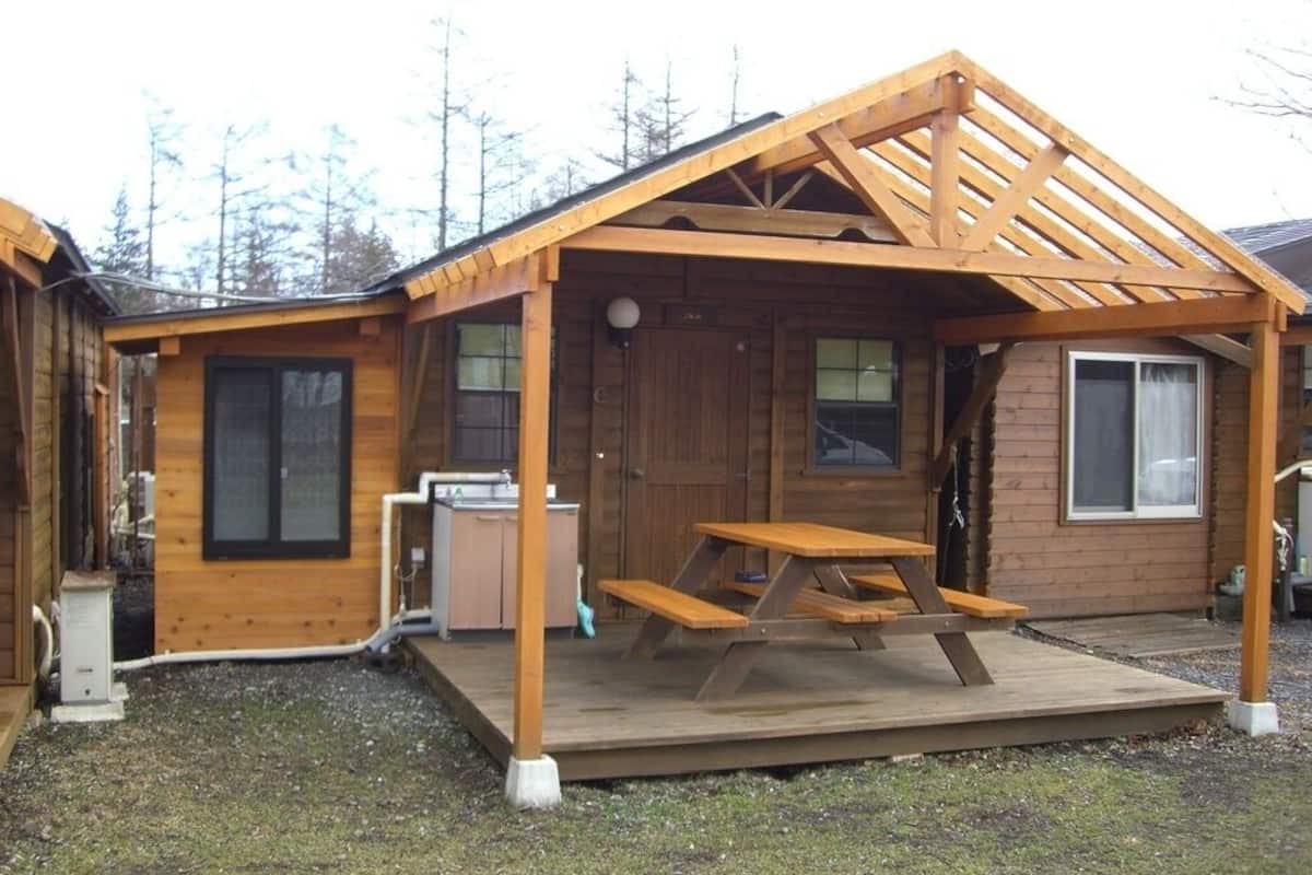 A wooden cabin with a covered porch, a picnic table, and a small kitchen area.