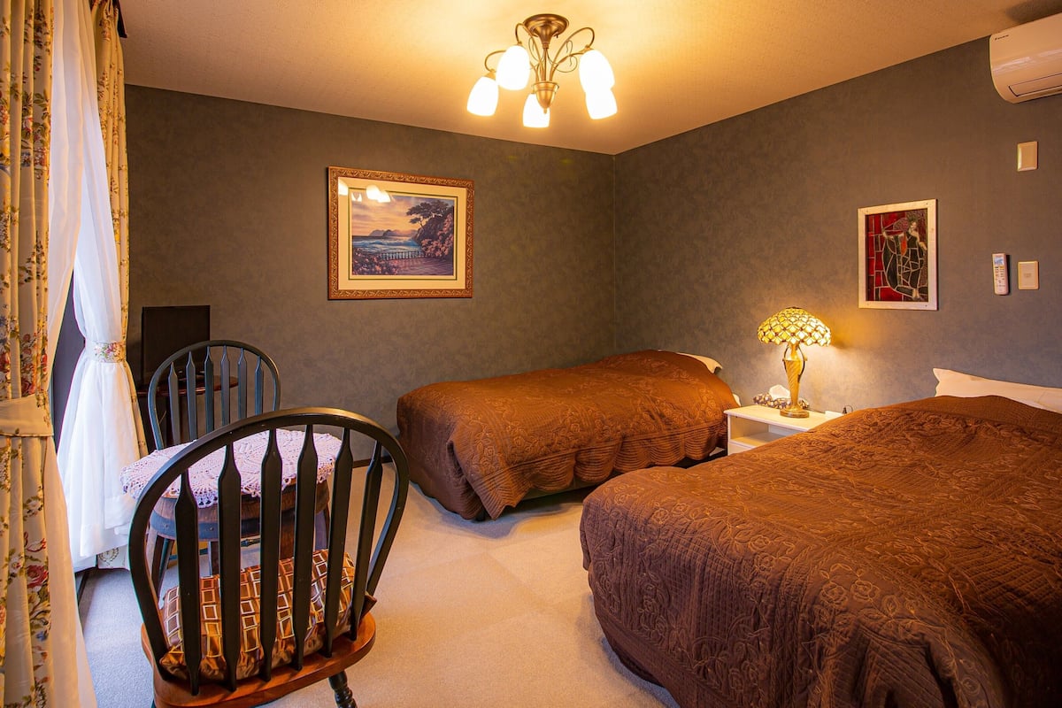 A room with two beds, a chair, a table with a lamp, a framed picture on the wall, and an air conditioner.
