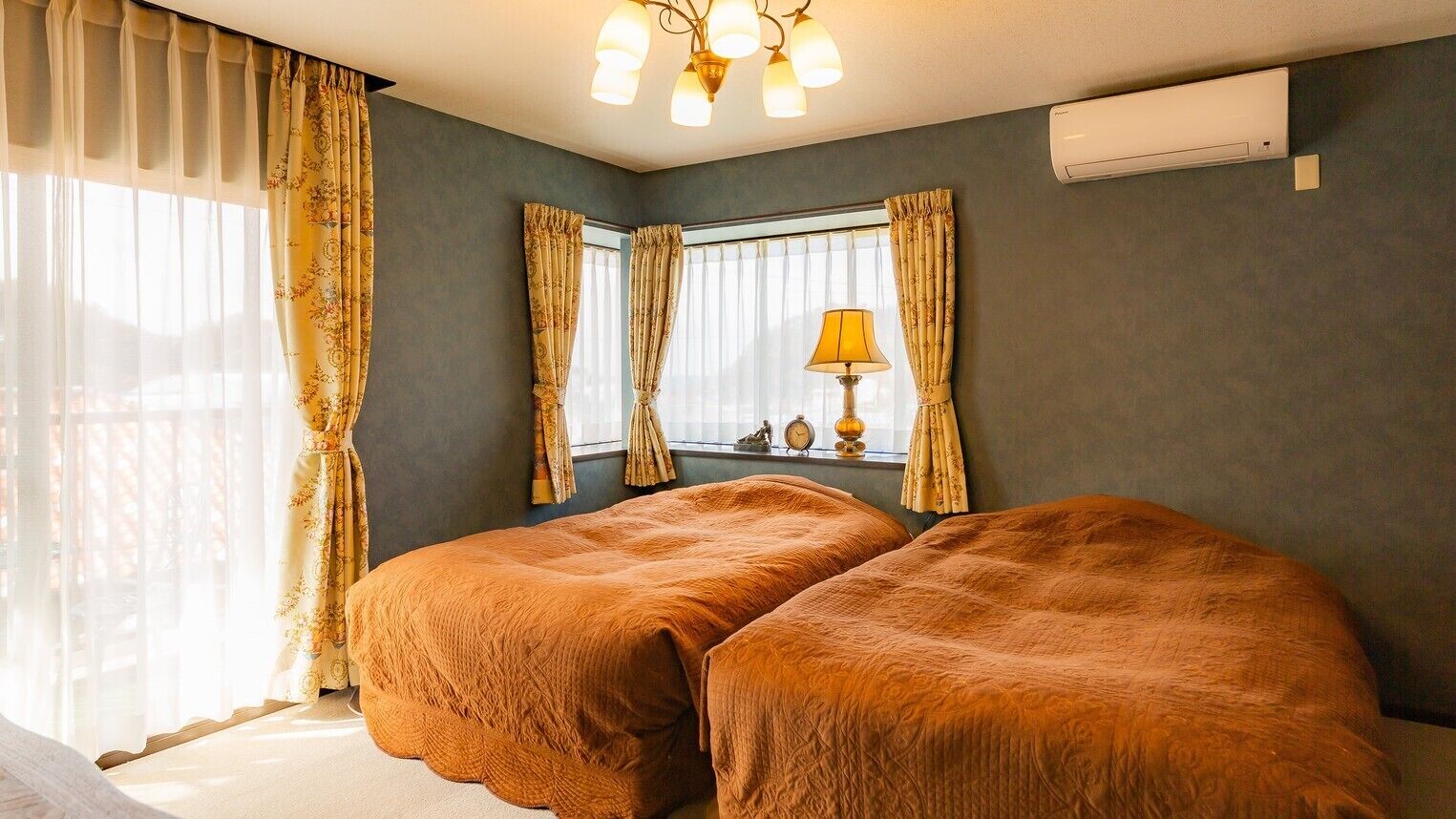 Ein Zimmer mit zwei Betten, einem Fenster mit Vorhängen, einer Lampe und einer Klimaanlage.
