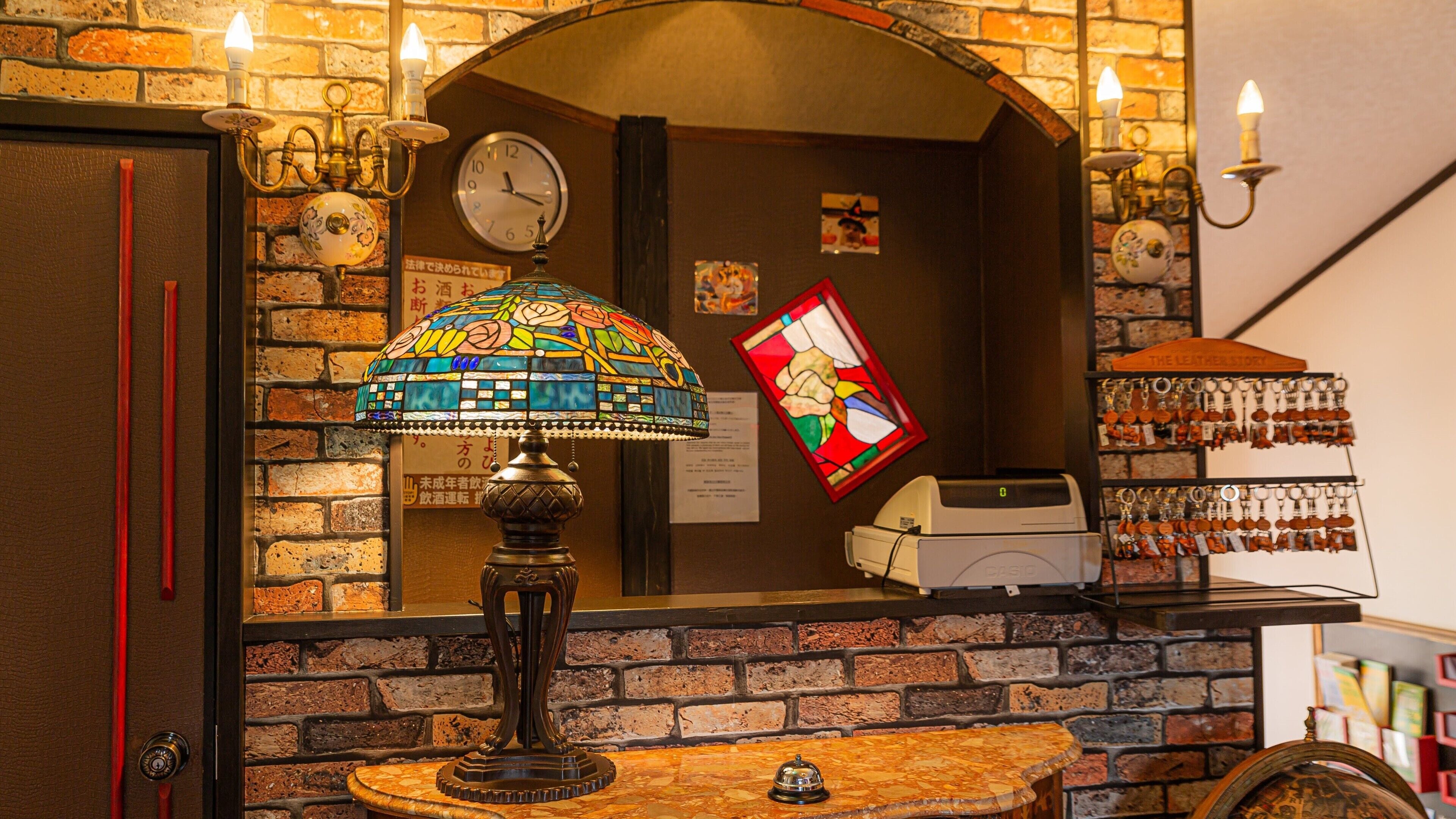 A hotel lobby with a brick wall, a clock, a stained glass lamp, a cash register, and a key display.