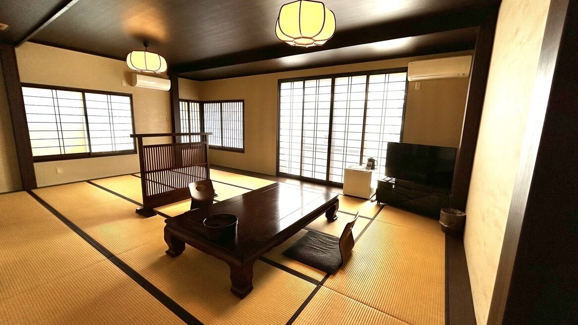 Ein Raum mit Tatami-Matten, einem niedrigen Tisch, einer Bank, einem Fernseher und Schiebetüren.