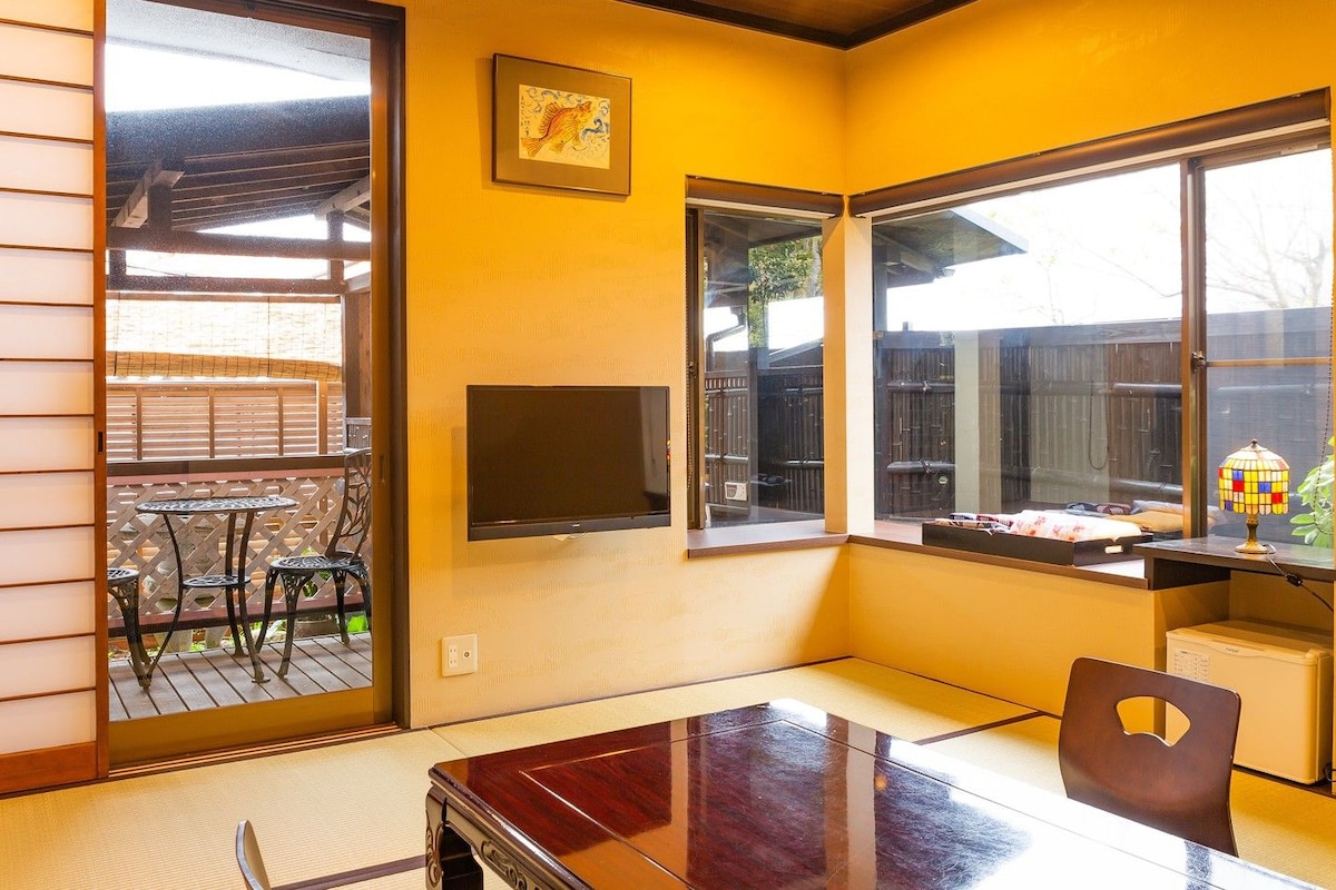 A room with tatami flooring, a sliding door leading to an outdoor area, a TV mounted on the wall, and a table with chairs.