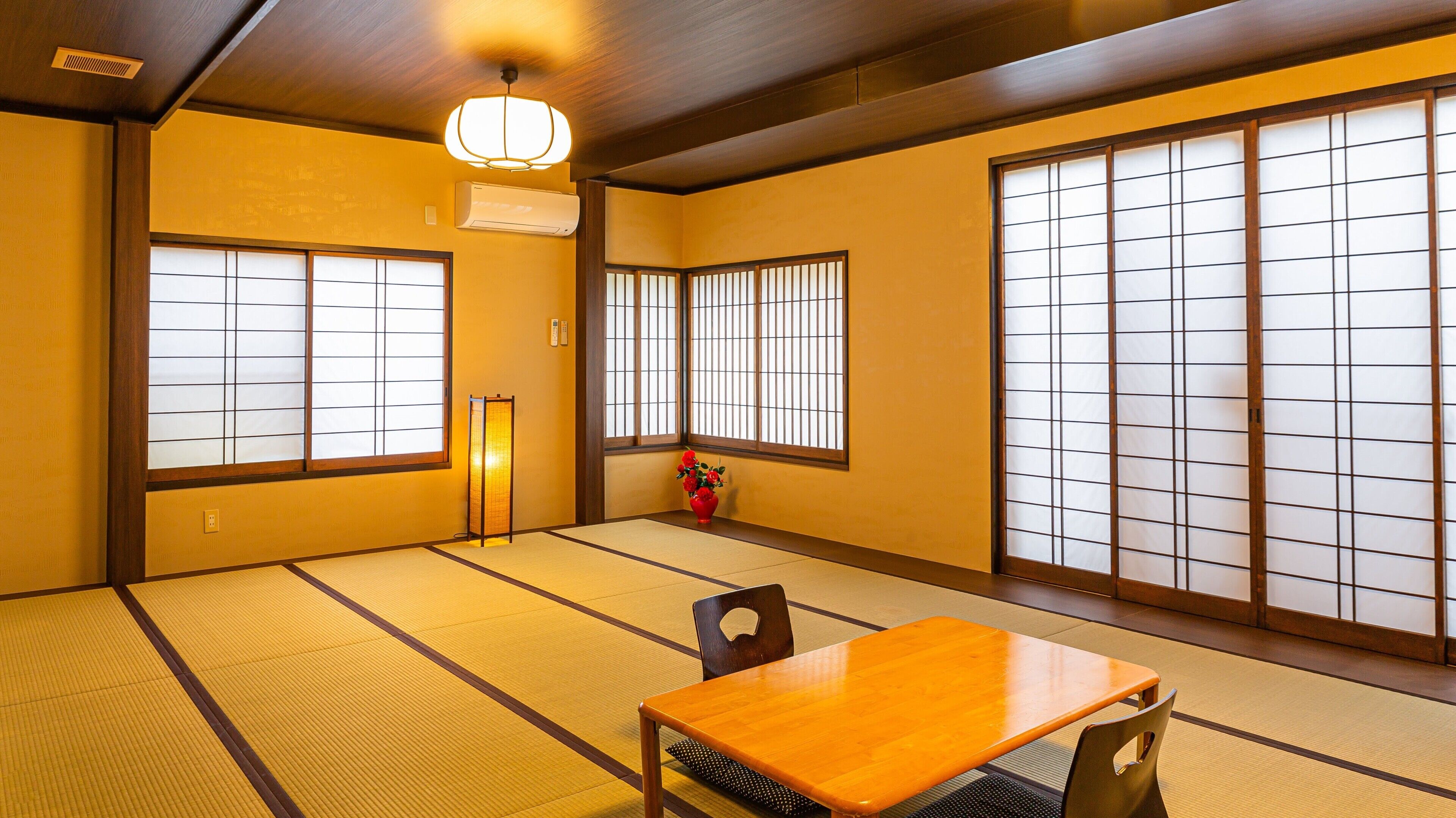 Ein traditioneller japanischer Raum mit Tatami-Boden, Shoji-Schirmwänden, einem Holztisch und einer roten Vase mit Blumen.