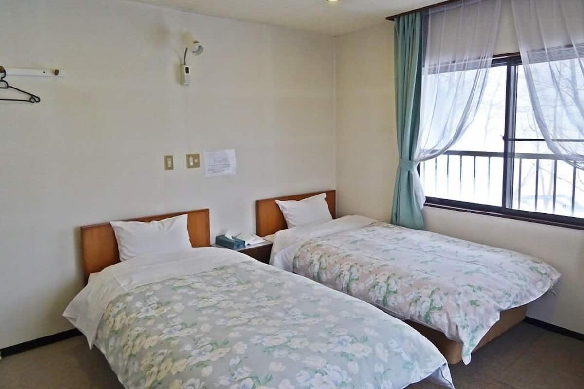 Habitación de hotel con dos camas, una ventana con cortinas translúcidas, una lámpara de pared y un aviso en la pared.