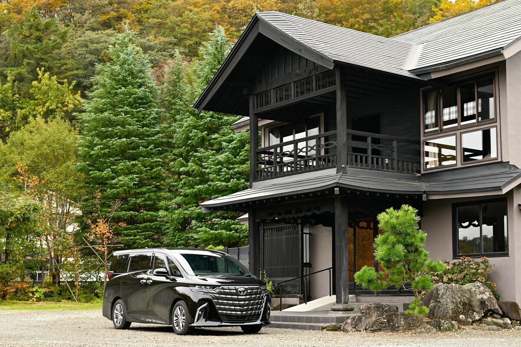 Un bâtiment à deux étages doté d’un balcon, avec une voiture garée devant et une forêt en arrière-plan.