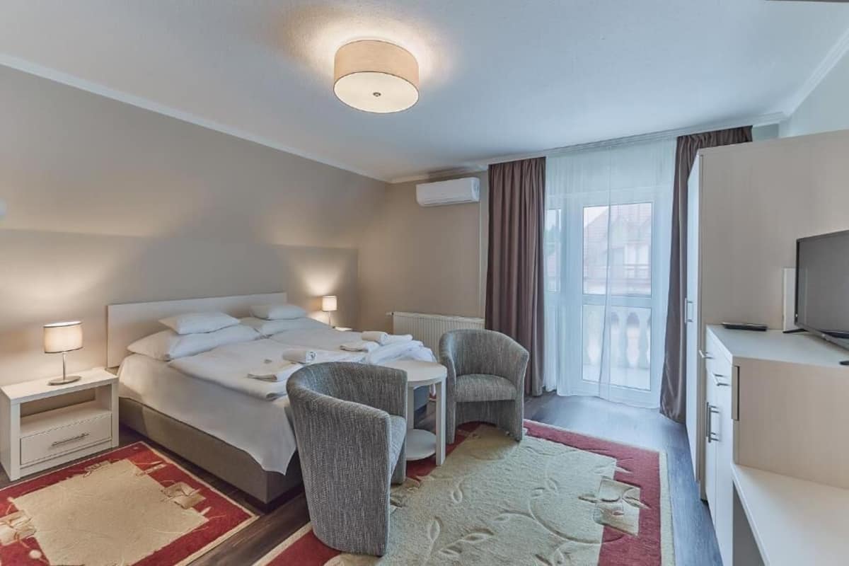 Une chambre d’hôtel avec un lit, deux fauteuils, une petite table, un téléviseur et une fenêtre avec des rideaux.