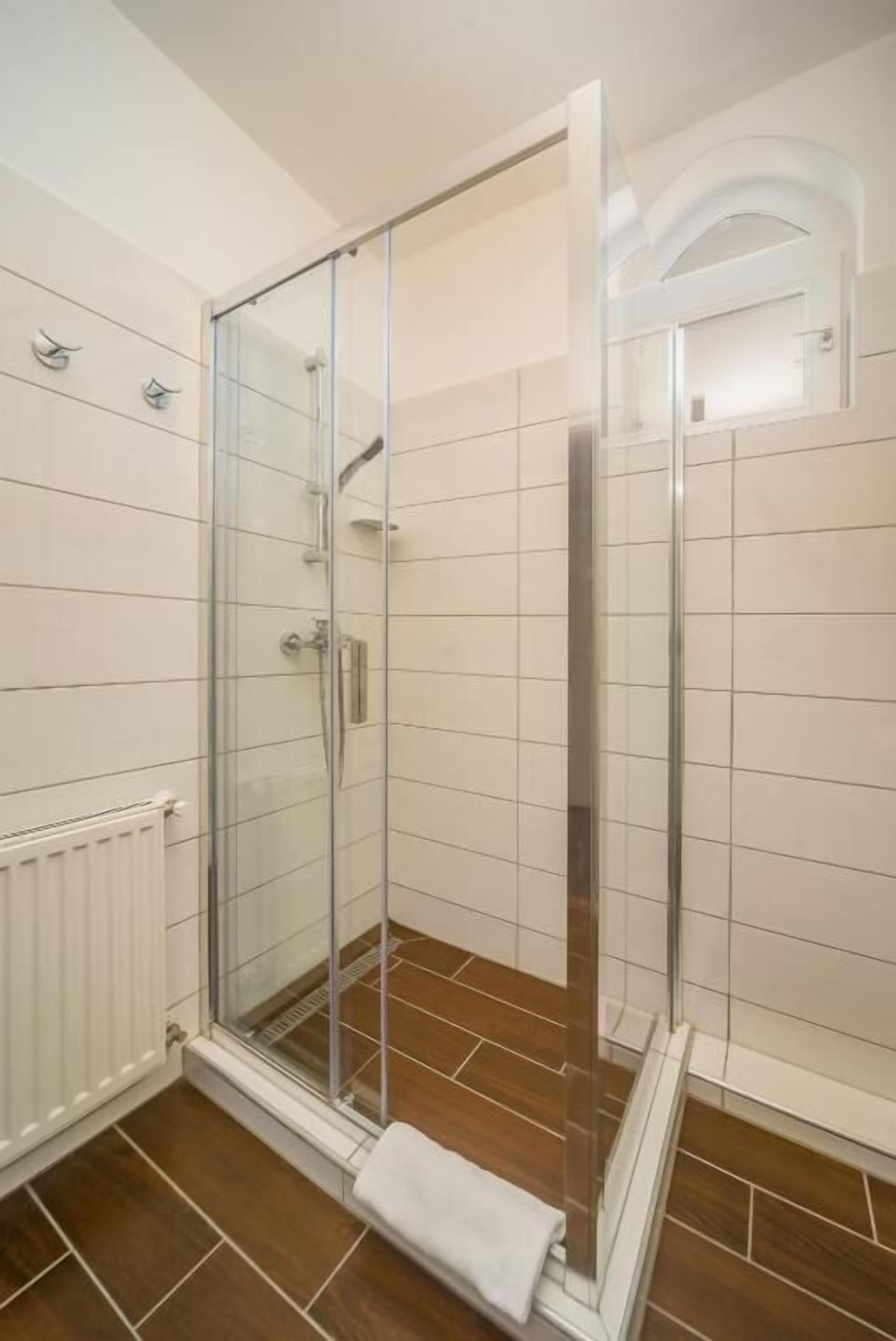 A glass-enclosed shower with a white towel on the floor, white tiled walls, and a wooden floor.