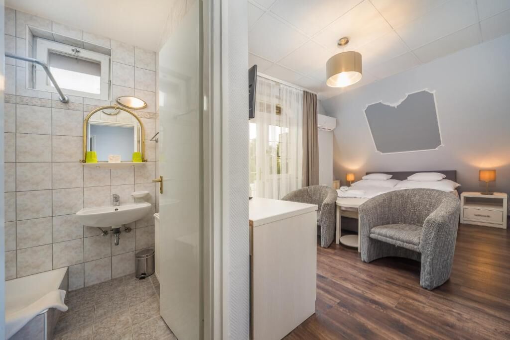 A modern bathroom with a sink, mirror, and shower. The room has a bed with a headboard, bedside tables, and a chair.