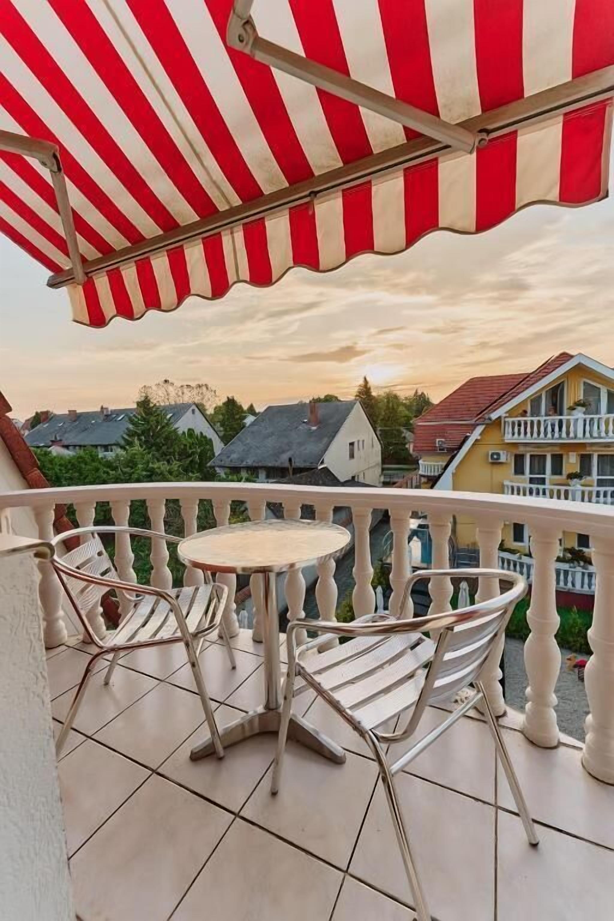 Ein Balkon mit gestreifter Markise, zwei Stühle, ein kleiner runder Tisch und Blick auf Häuser im Hintergrund.