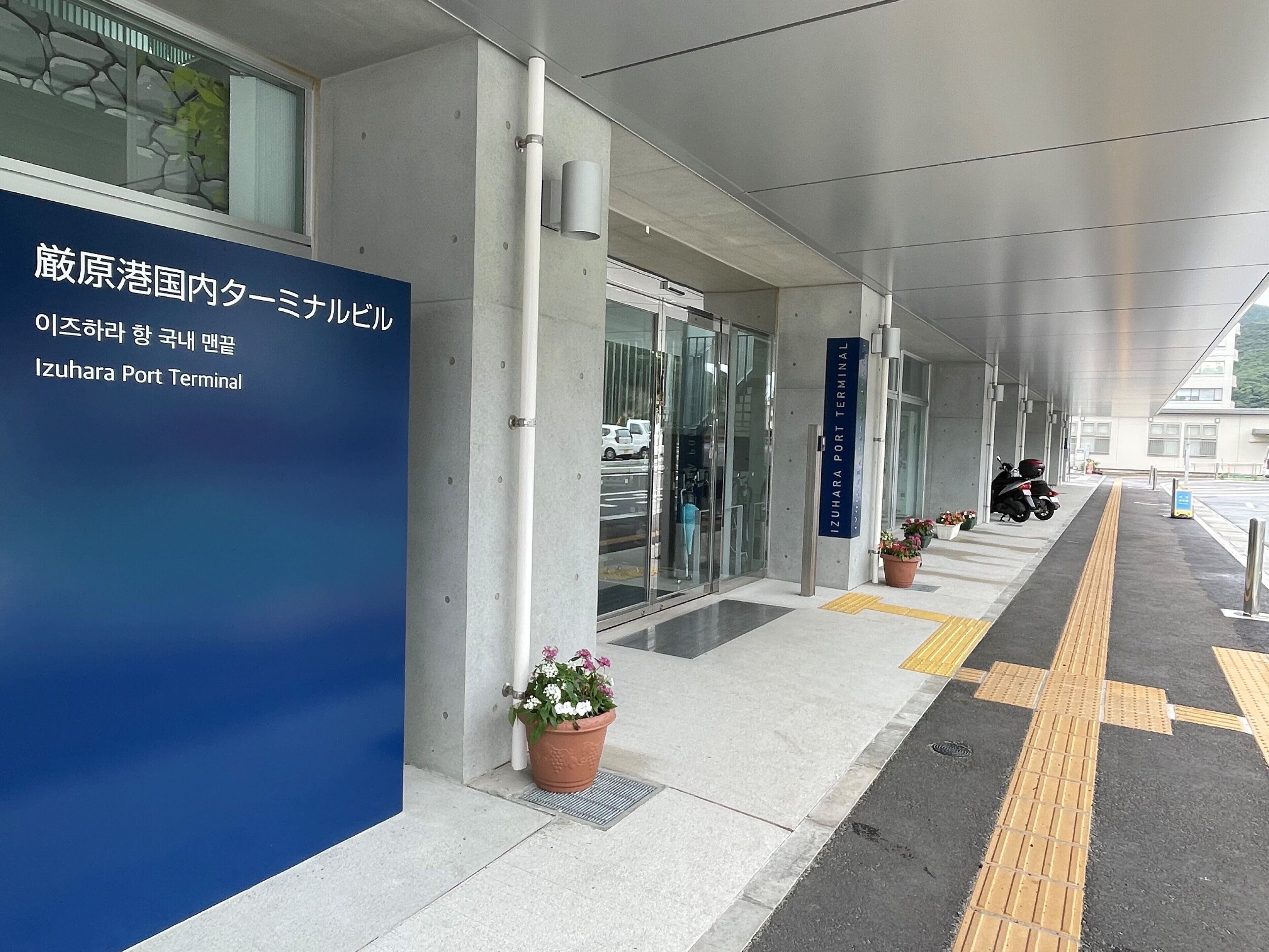 A covered walkway at Izuhara Port Terminal with a blue sign, potted plants, and tactile paving for the visually impaired.