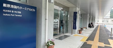 Une galerie couverte au terminal portuaire d’Izuhara, agrémentée d’un panneau bleu, de plantes en pot et d’un revêtement de sol tactile pour les personnes malvoyantes.