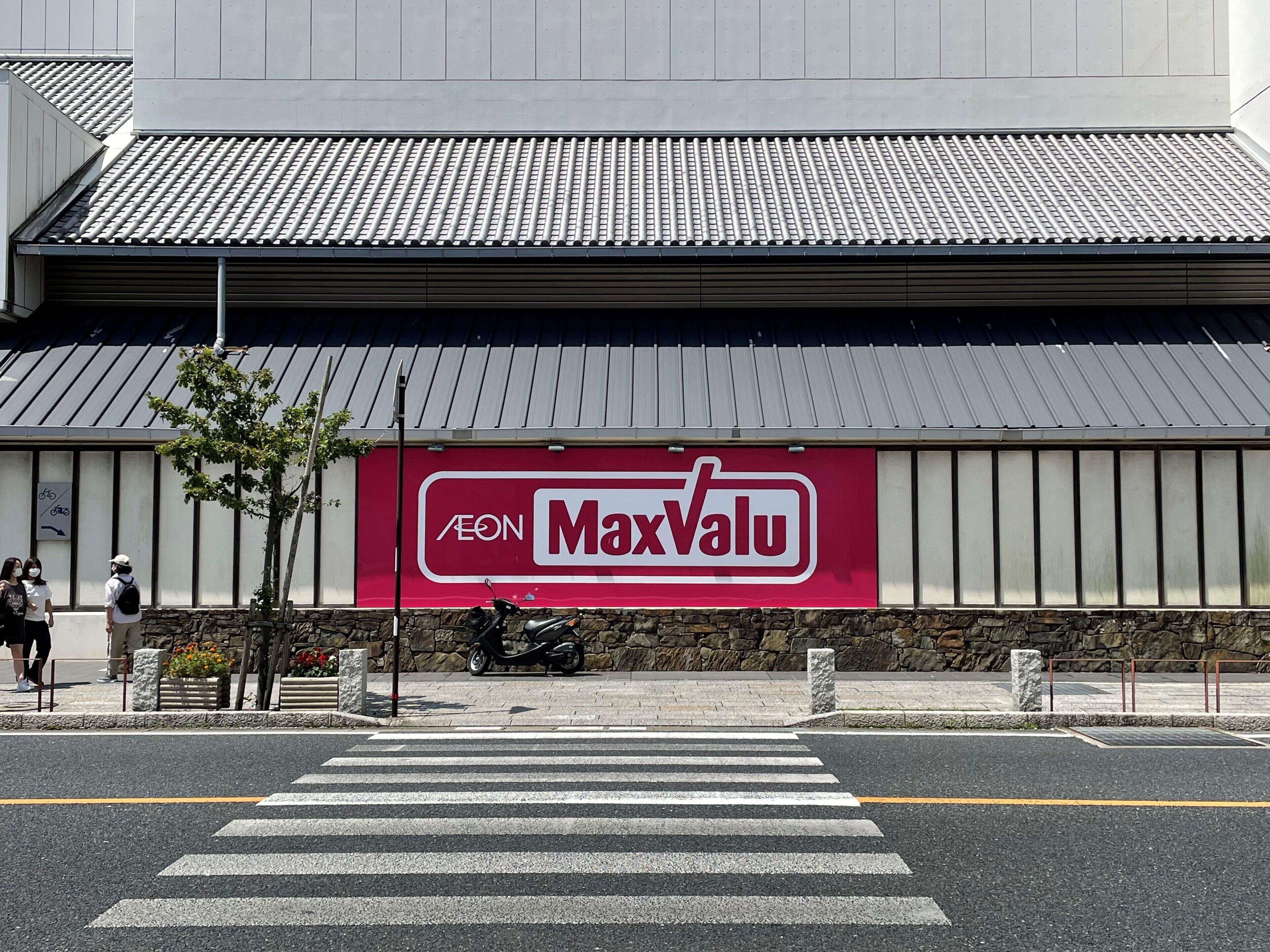 Een gebouw met een grote MaxValu reclamebord, een zebrapad en een geparkeerde scooter.