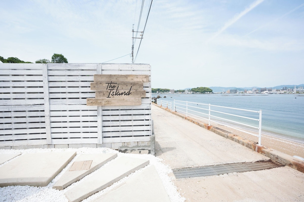 Un paseo marítimo con un letrero que dice 'The Island', una cerca blanca, escalones de hormigón y una barandilla metálica a la orilla del agua.