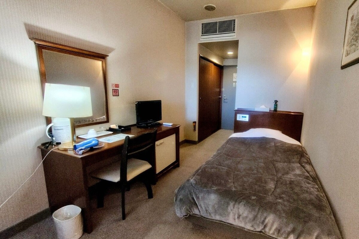 A hotel room with a bed, a desk with a computer, a chair, a lamp, and a mirror.