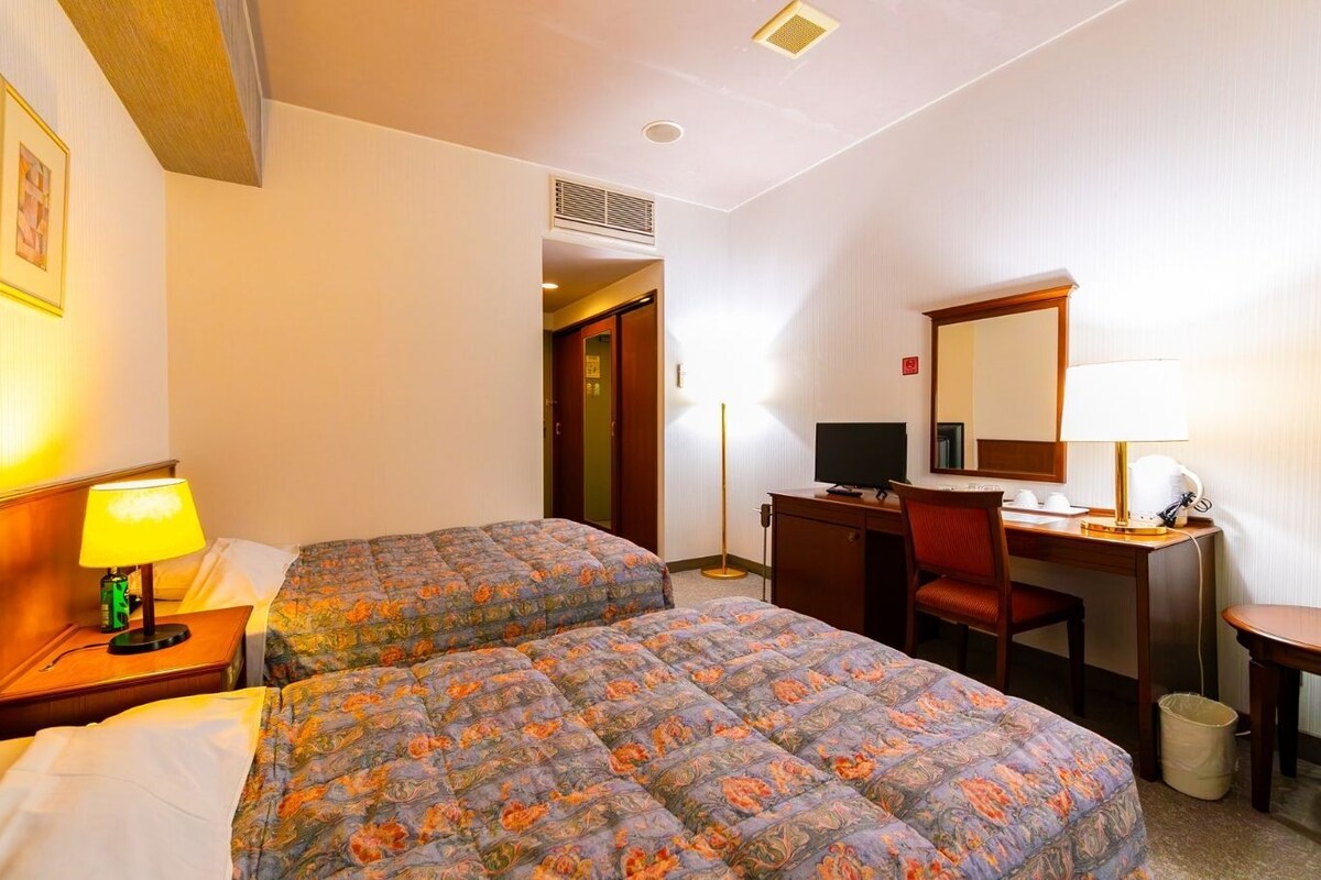 A hotel room with two beds, a desk, a chair, a computer, and a lamp.