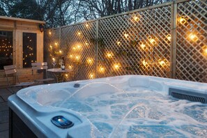 A hot tub with a control panel, surrounded by a wooden fence with string lights, and a wooden cabin with a table and chairs. - Luxury Family Period Home By The Cotswold Way (Leonard Stanley)
