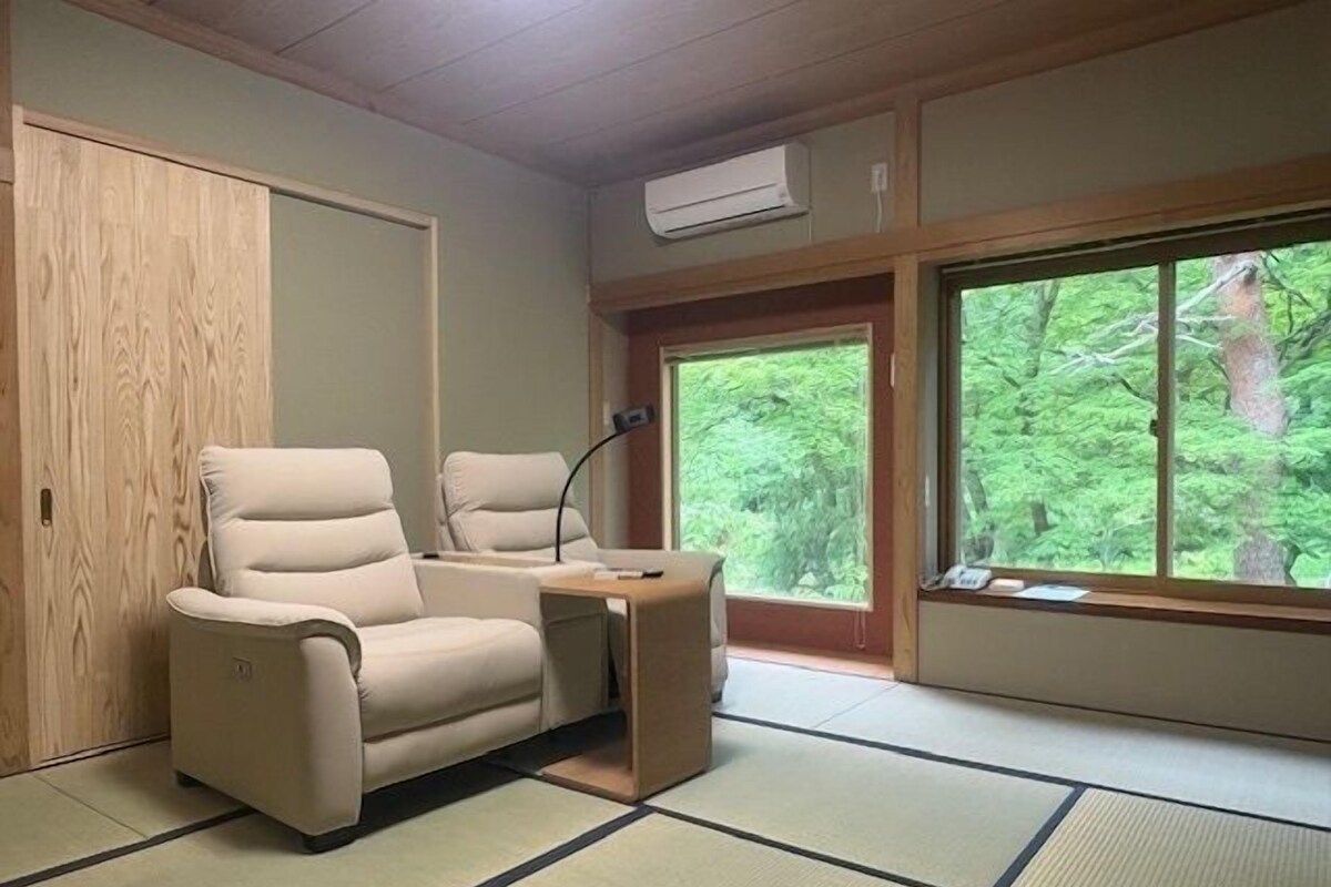 A room with tatami flooring, a sofa, a desk, and large windows with a view of trees.