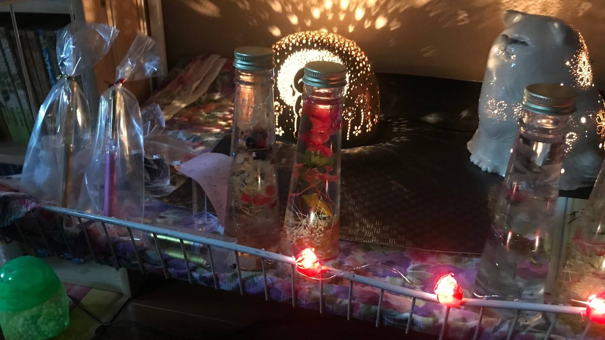 Egy polc, amelyen különböző tárgyakat tartalmazó üvegek találhatók, egy díszes macskafigurina és egy sor fényfüzér.