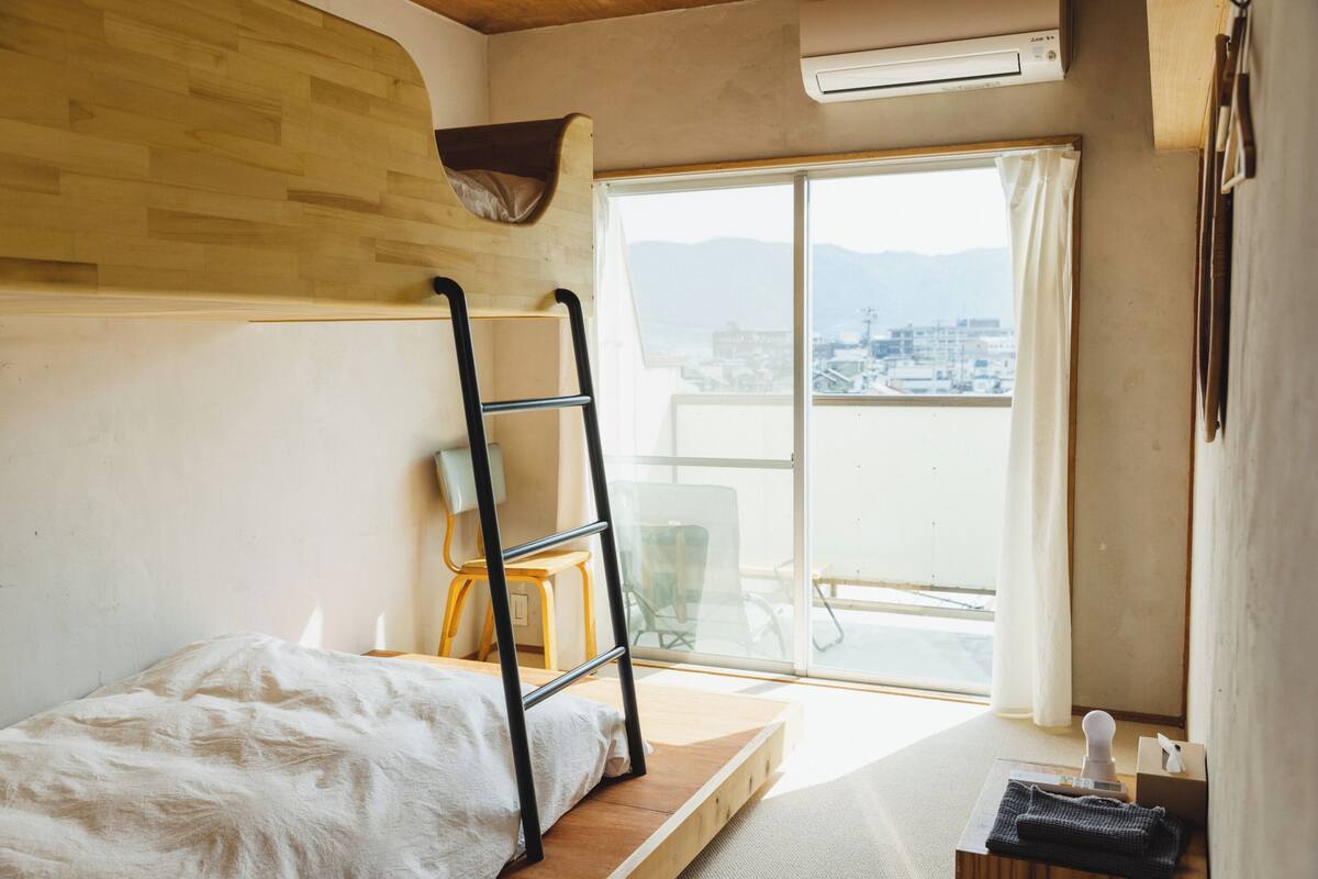 A room with a bunk bed, a ladder, a window with a view of buildings and mountains, and a small table with a chair.