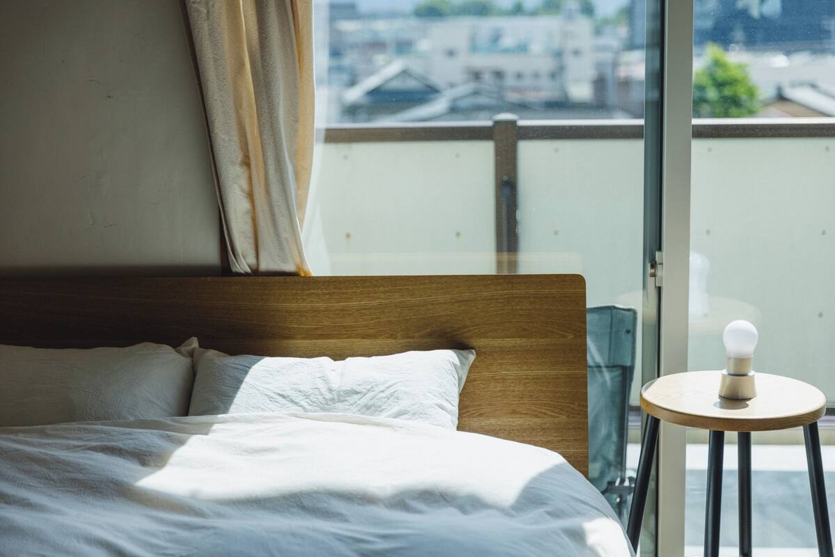 A hotel room with a bed, a window with a view of buildings, and a small table with a light bulb.