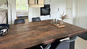 Uma sala de jantar com mesa de madeira, cadeiras, luminária pendente, televisão e janela com persianas.