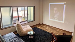 Una habitación con sofá, sillón, una mesita, pantalla de proyección y un ventanal grande.