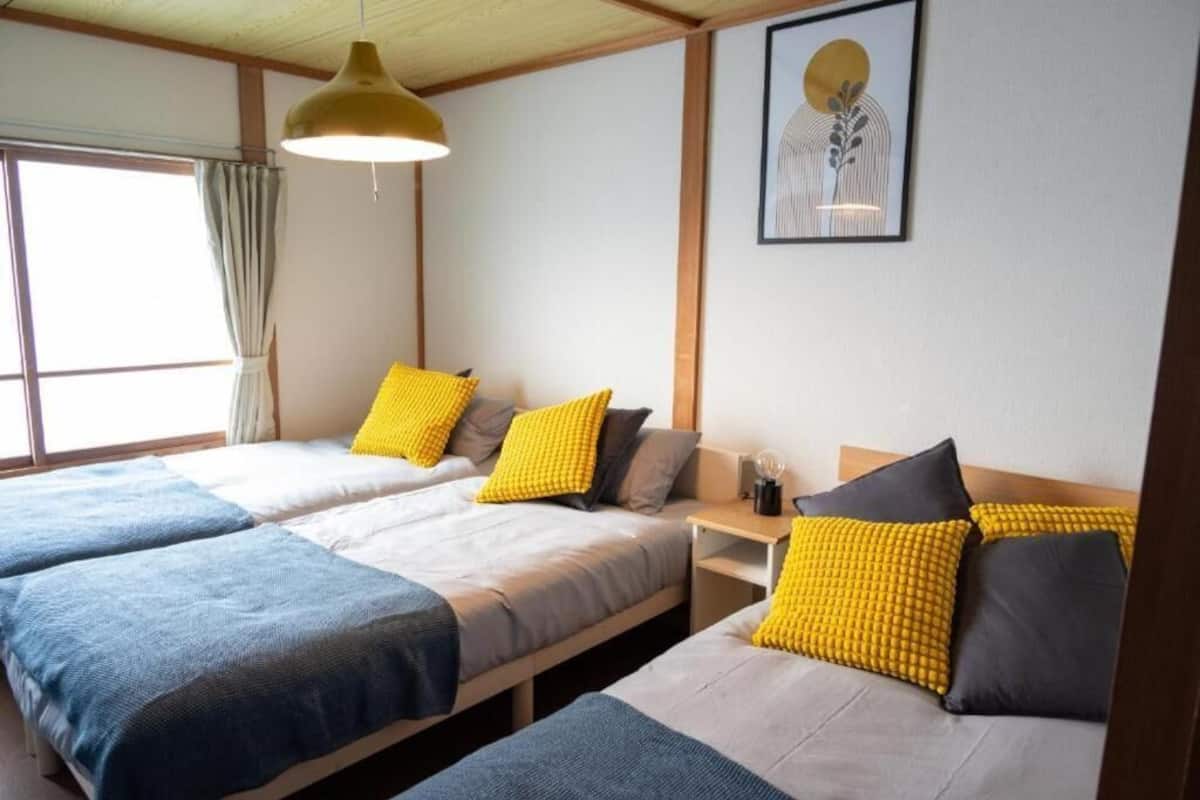 Ein Zimmer mit zwei Betten, jeweils mit einer blauen und grauen Bettdecke und gelben Kissen. An der Wand hängt ein gerahmtes Kunstwerk und ein Fenster mit Vorhängen.