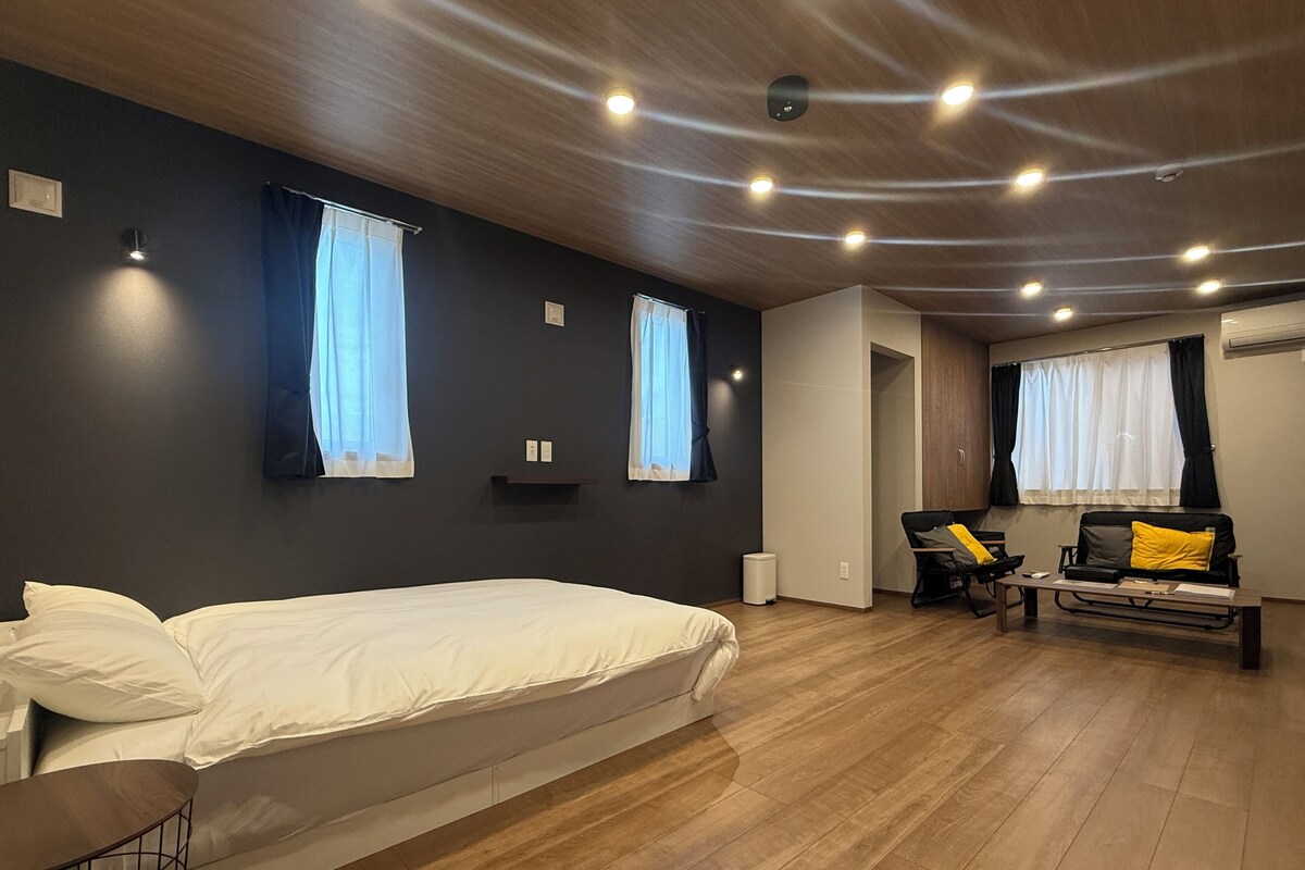 A modern hotel room with a large bed, a sofa, a small table, and a desk. There are two windows with curtains, a ceiling with recessed lighting, and a wooden floor.