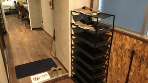 A hotel room with a wooden floor, a black shoe rack with slippers, a mat with a shoe-off sign, and a bed in the background.