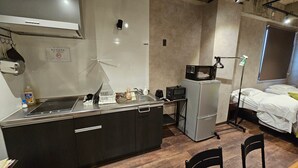 A compact kitchen with a microwave, sink, and stove, a small refrigerator, and a bed with white bedding.