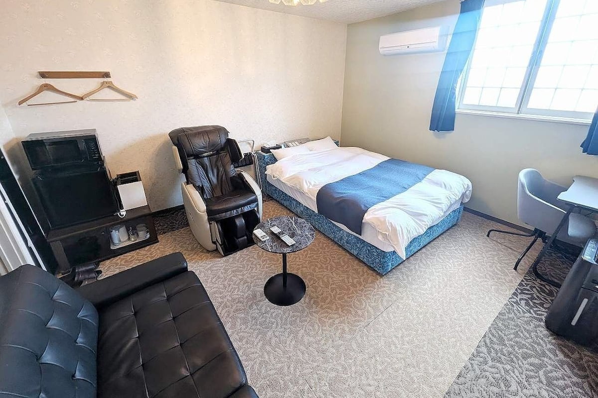 A hotel room with a bed, a massage chair, a small table, a desk, and a TV.