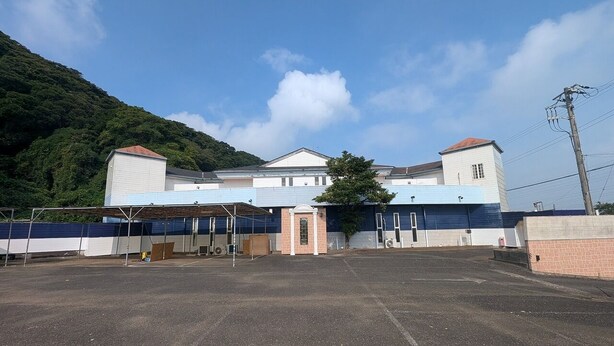 A large parking lot with a row of buildings, including a white building with a red roof and a blue building with a white roof. - Selfplus Hotel Choshi (Choshi)