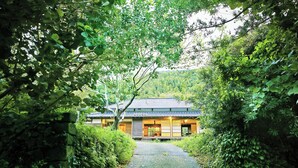 Una casa tradicional japonesa con techo de tejas, rodeada de abundante follaje y un camino de grava que conduce a la entrada.