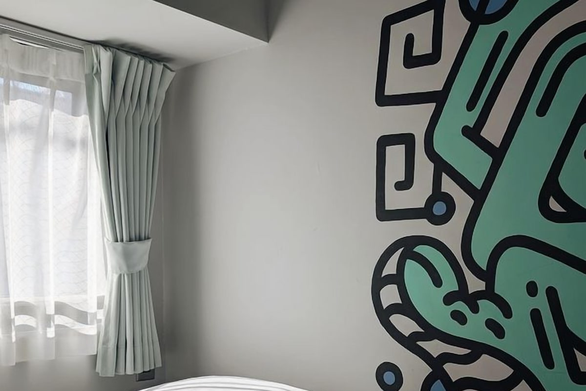 A bedroom with a bed, a window with curtains, and a wall with a geometric mural.