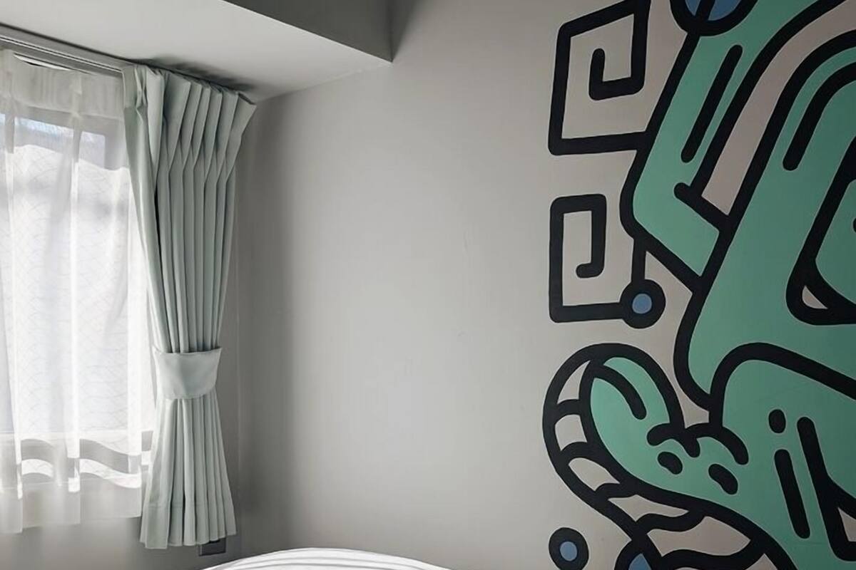 Une chambre avec un lit, une fenêtre avec des rideaux et un mur orné d’une fresque géométrique.