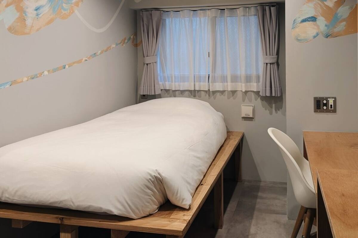 Un lit simple avec un cadre en bois, un petit bureau avec une chaise et une fenêtre ornée de rideaux légers.