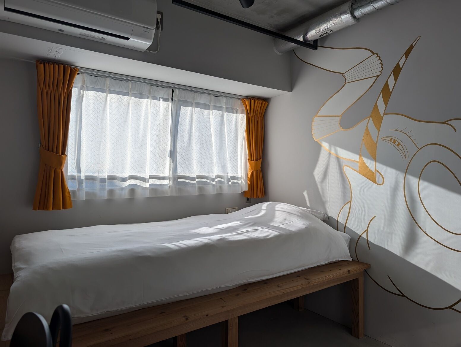 Ein Einzelbett mit Holzseitenverkleidung, ein Fenster mit durchsichtigen weißen Vorhängen und orangefarbenen Bändern sowie eine Wand mit einem großen abstrakten Gestaltungselement.