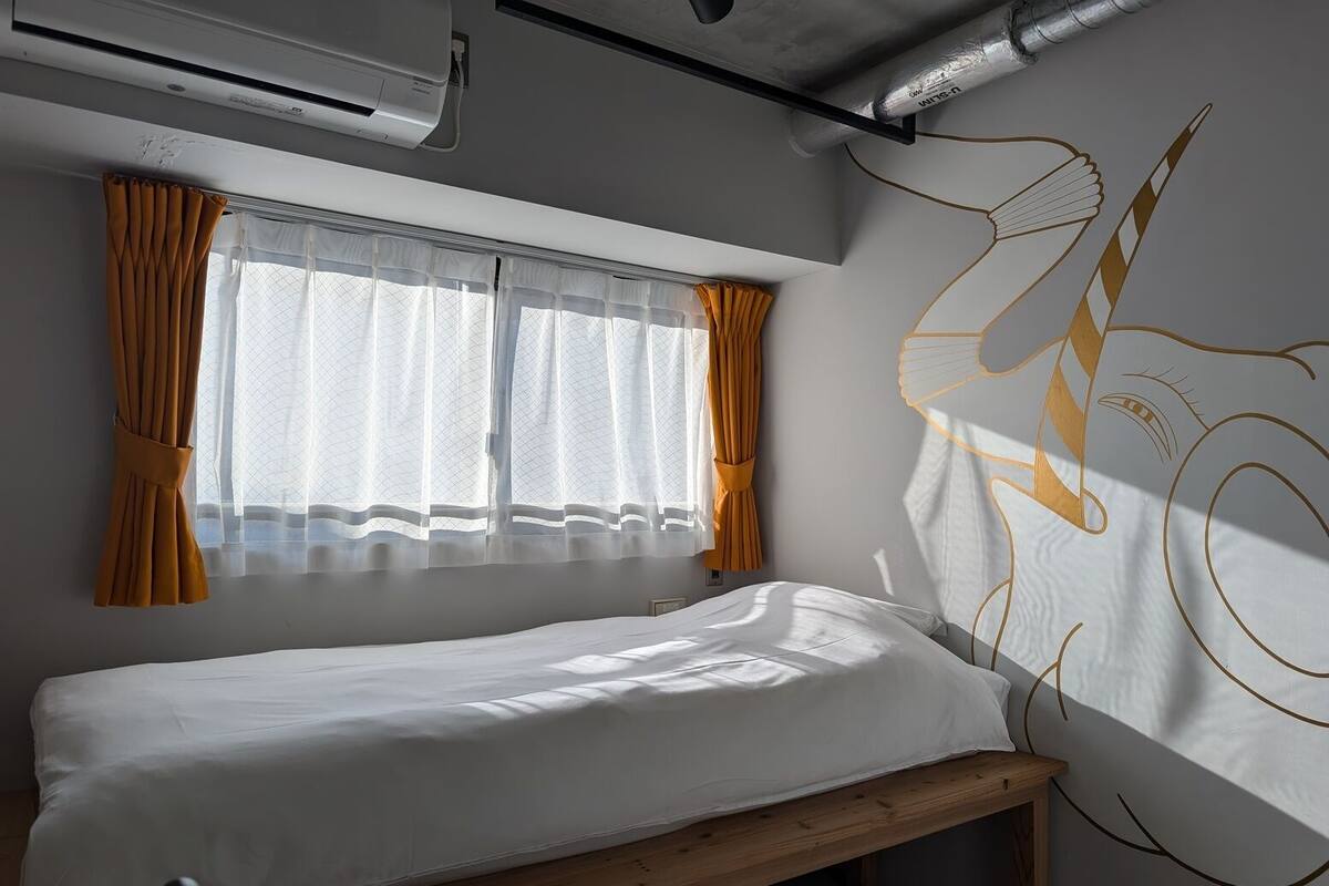 Un lit simple avec une tête de lit en bois, une fenêtre ornée de rideaux blancs légers et de liens orange, et un mur décoré d’un grand motif abstrait.