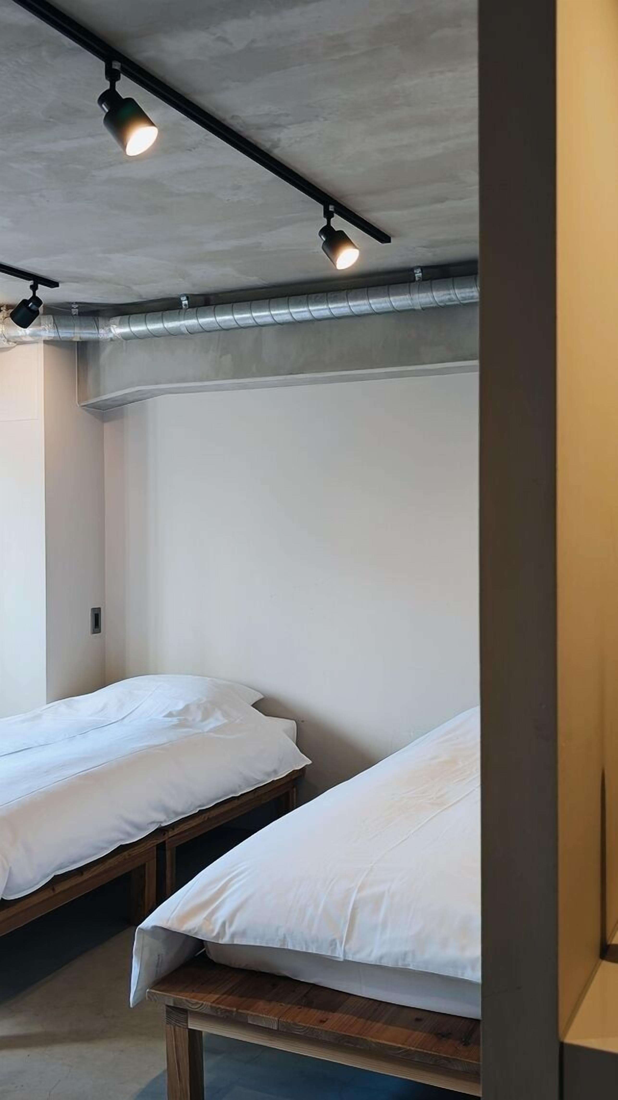 Ein Zimmer mit zwei Einzelbetten, einer Beton Decke und freiliegenden Leitungen.