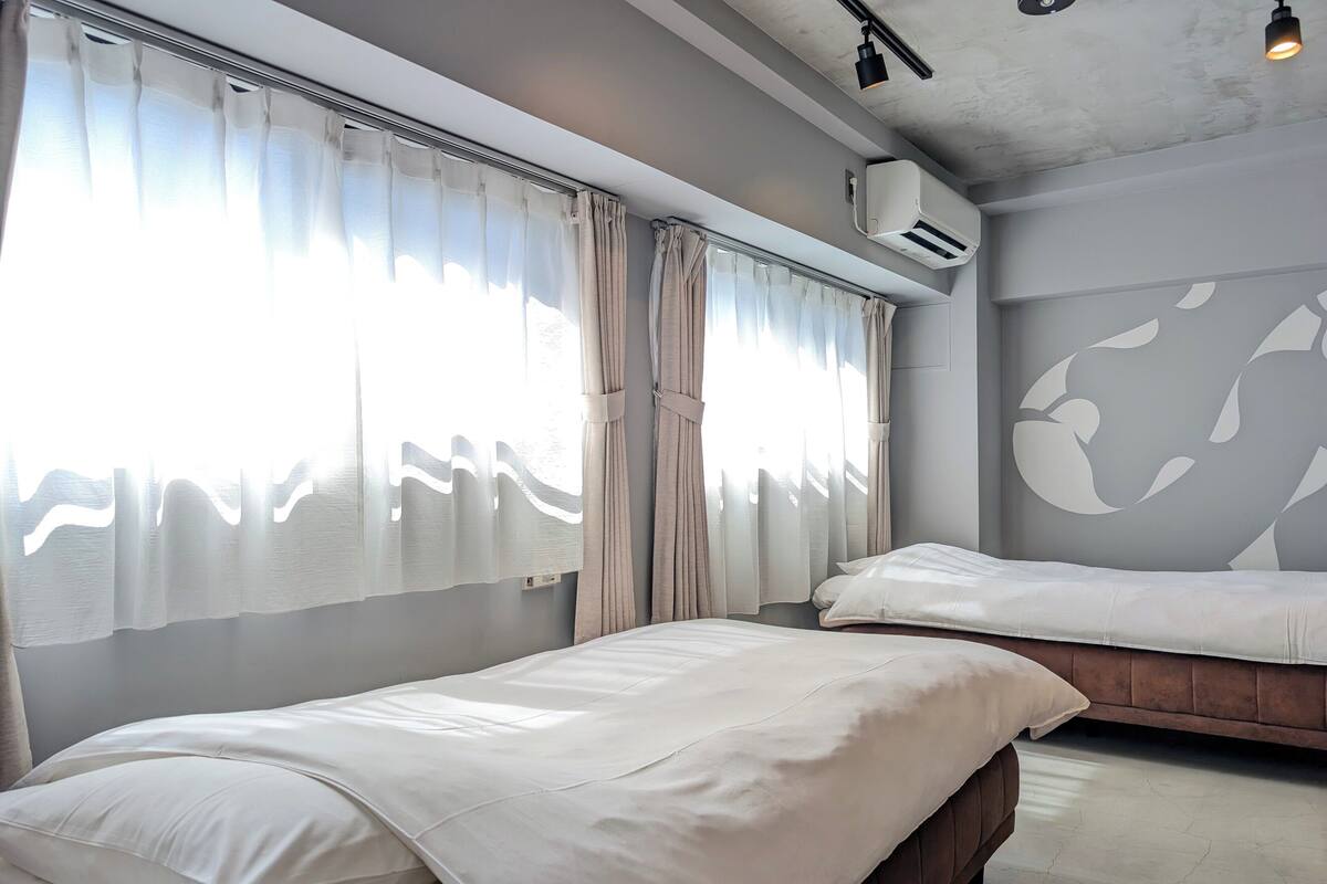 Une chambre d’hôtel avec deux lits, une grande fenêtre avec des rideaux légers, un appareil de climatisation et un mur orné d’un grand motif abstrait.