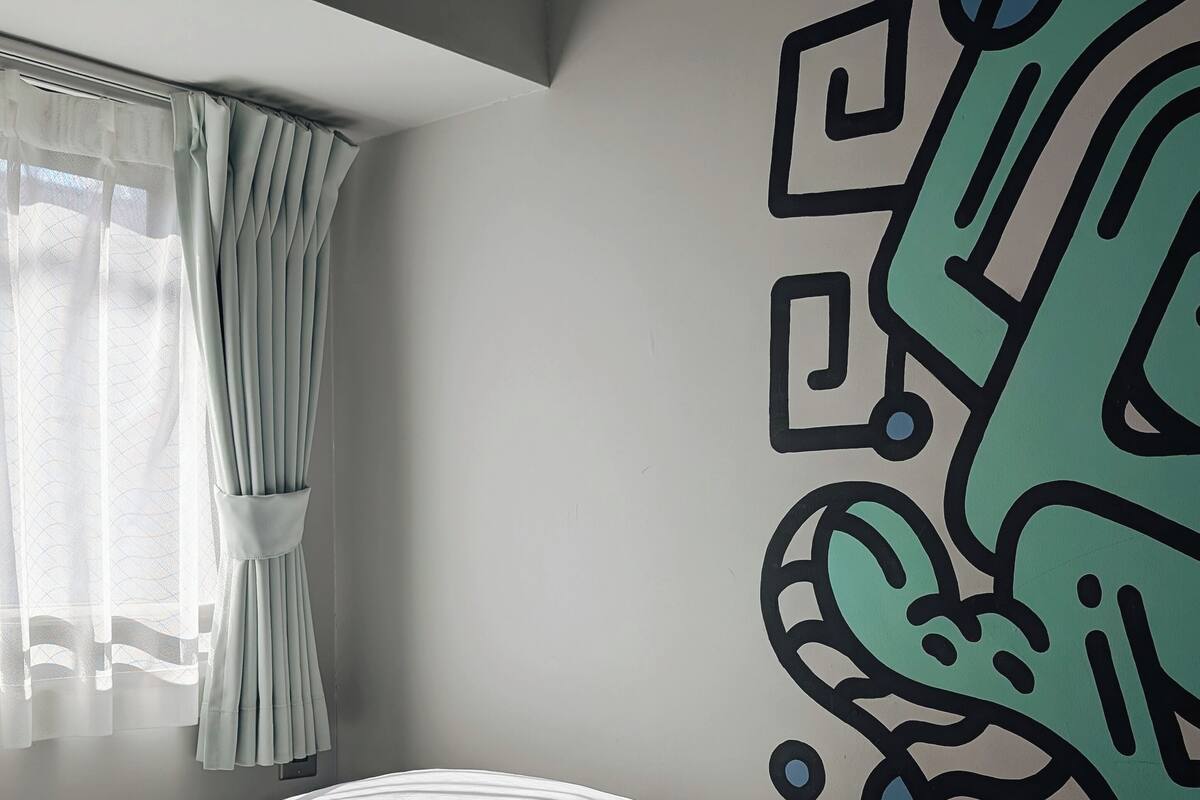 Une chambre à coucher avec un lit, une fenêtre avec des rideaux et un mur orné d’une fresque géométrique.